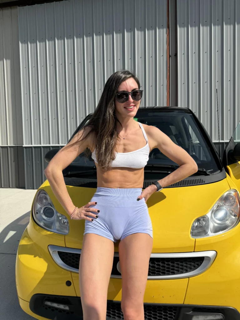 Bryce Adams (@fitbryceadams) Fansly model picture with car behind her. she is wearing white top and blue shorts